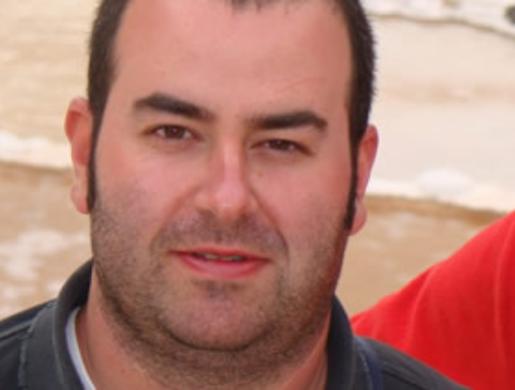 Uomo con barba corta, capelli scuri, sfondo di spiaggia e mare.