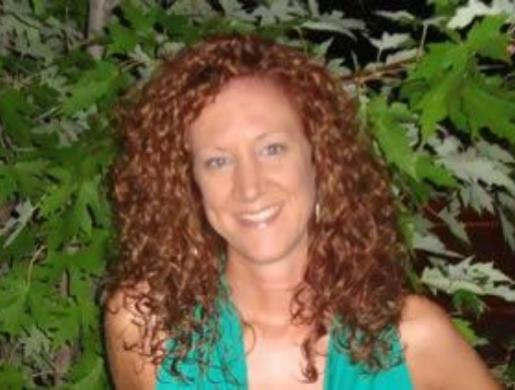 Donna sorridente con capelli ricci rossi, in piedi davanti a foglie verdi.