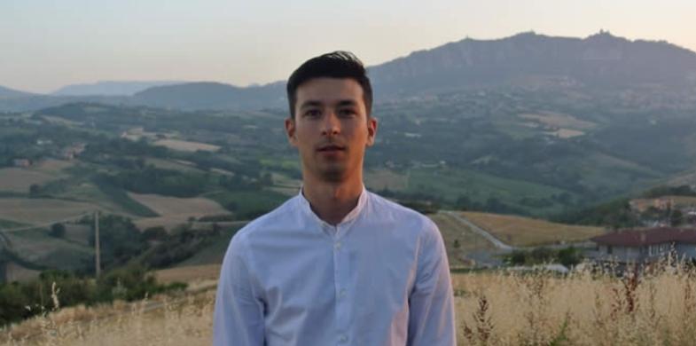 Giovane uomo in camicia bianca con sfondo di colline e paesaggio rurale.